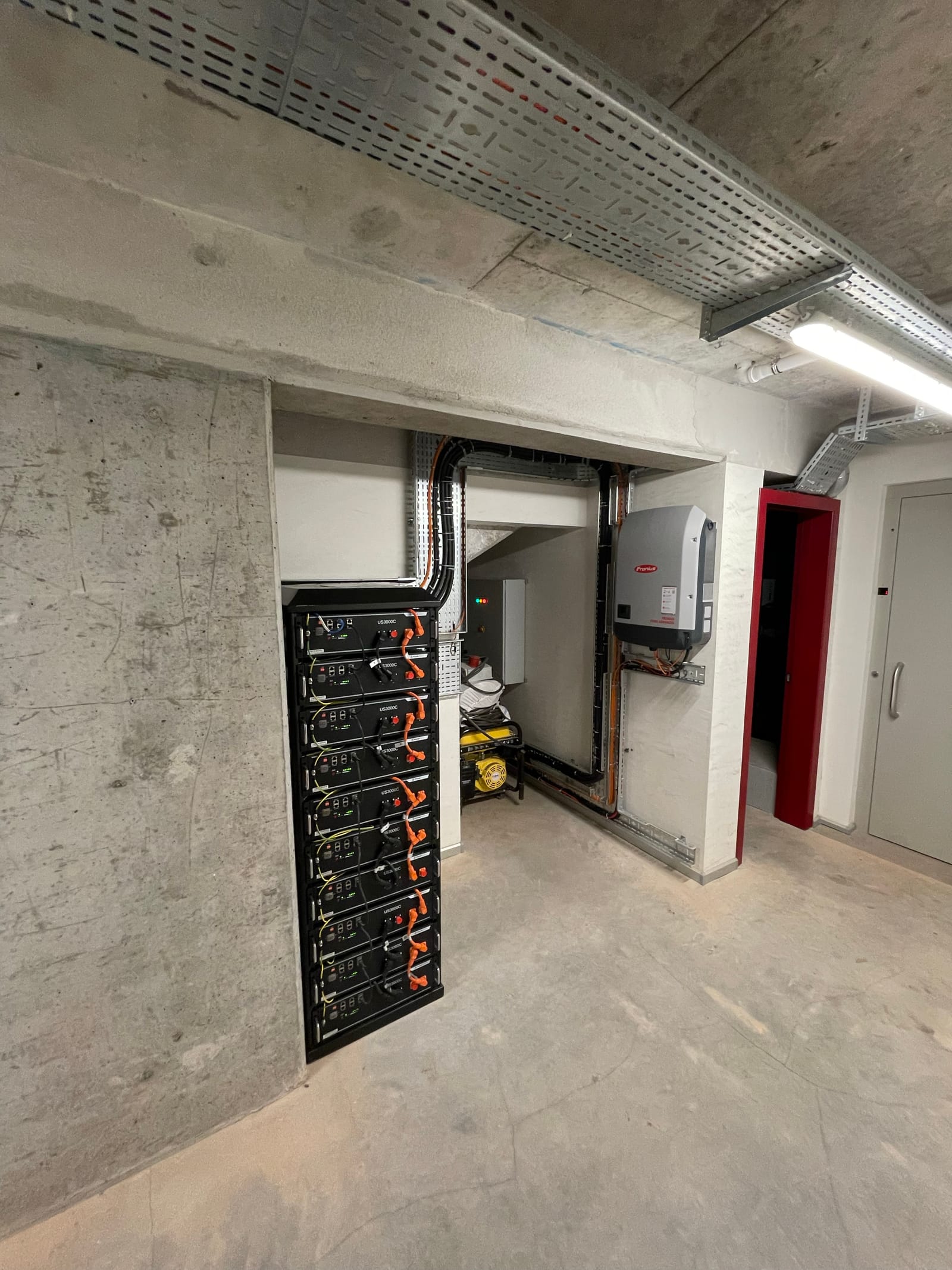 A rack of black battery modules and inverters in a bare concrete plant room.