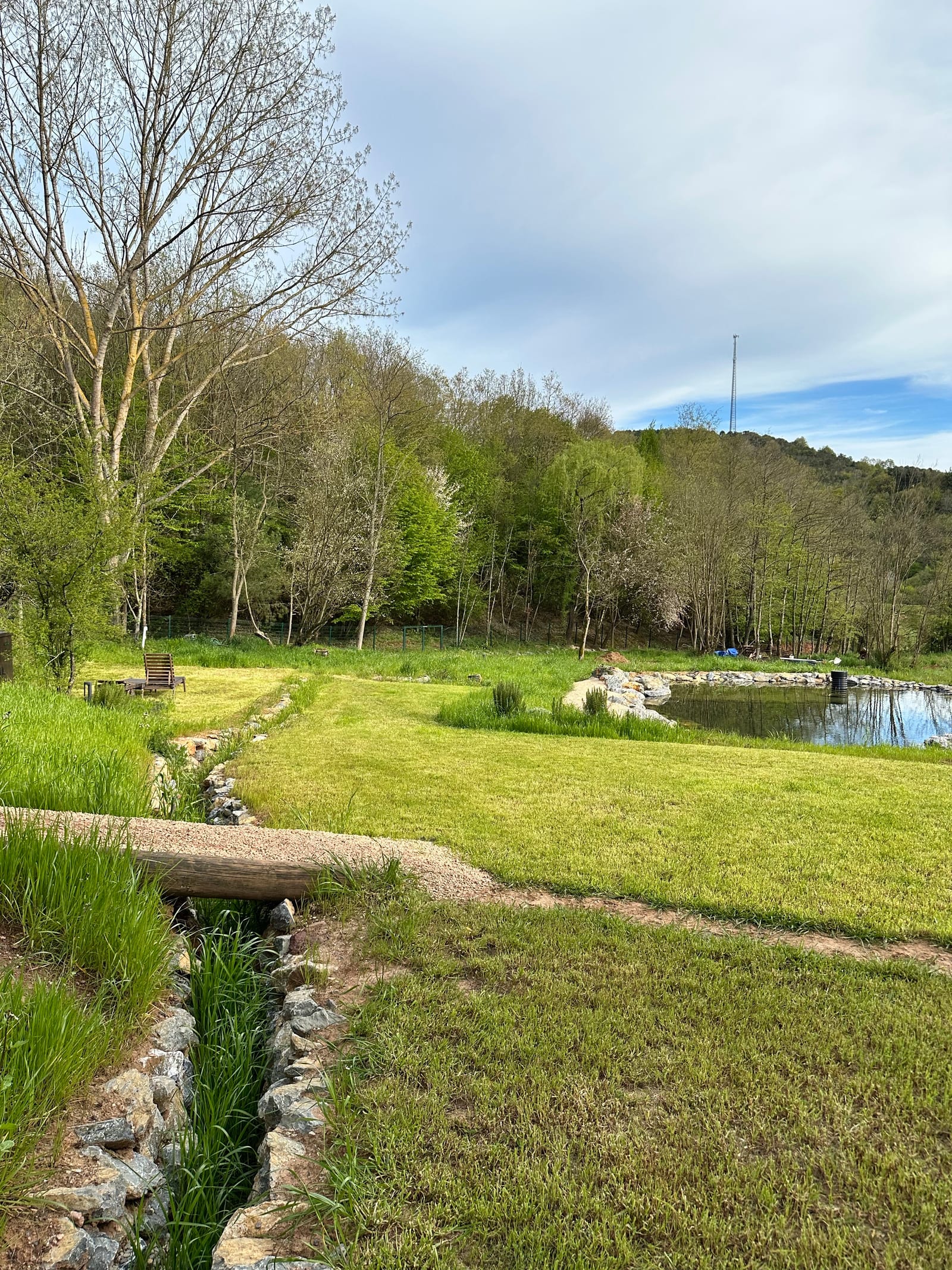A stone-lined channel running through grass toward a pond, with trees beyond.