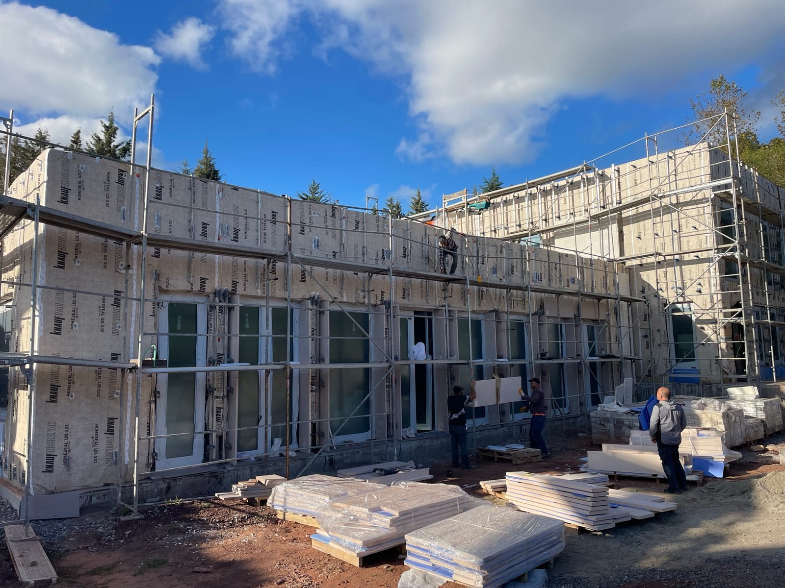 A building under construction wrapped entirely in pale mineral wool insulation, with scaffolding and workers.