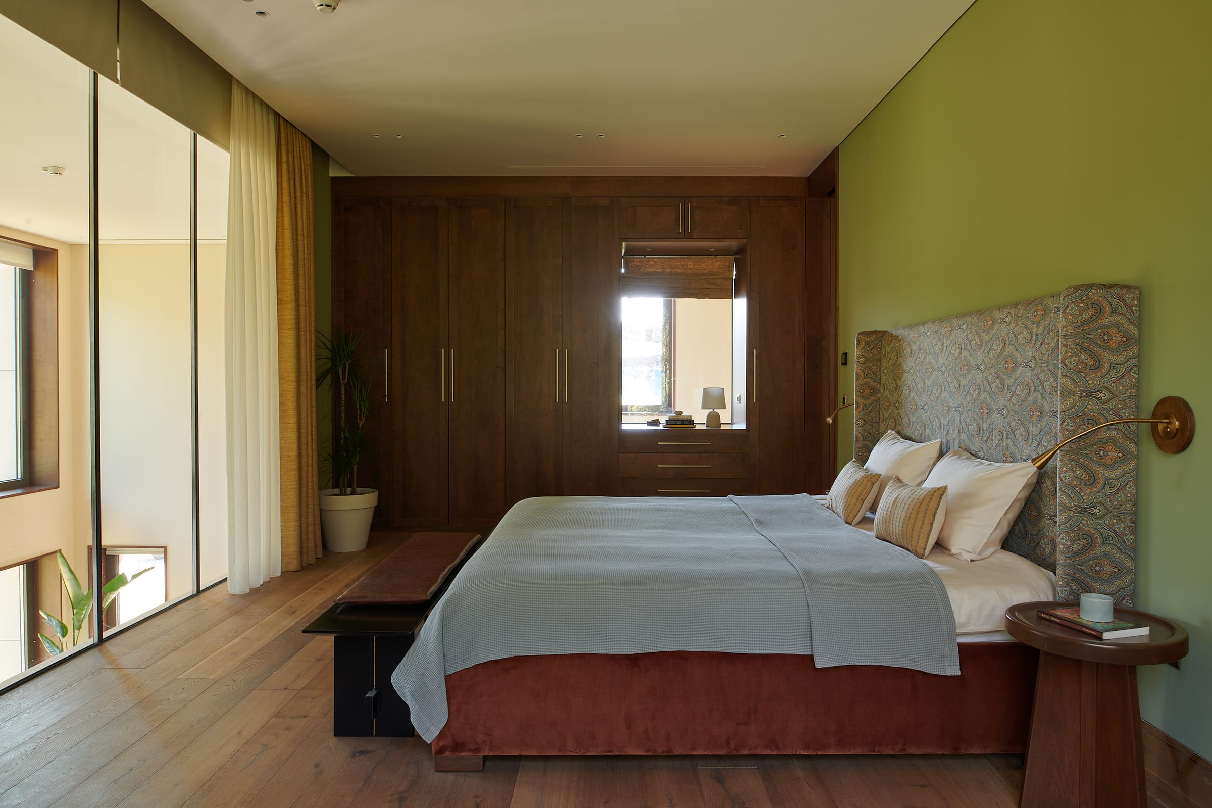 A bedroom with olive-green walls, a full-height timber wardrobe wall and a window onto open landscape.