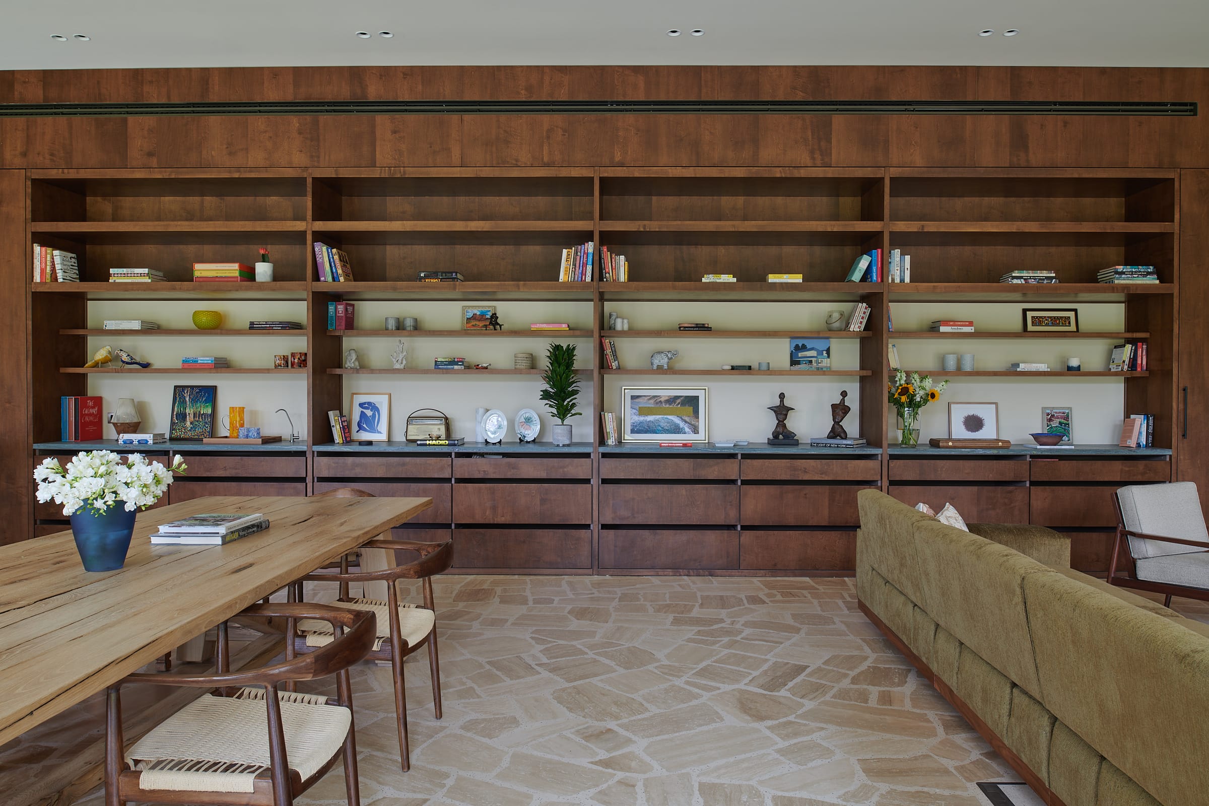 A living and dining space with a stone floor and a long timber shelving wall, opening through to a kitchen.