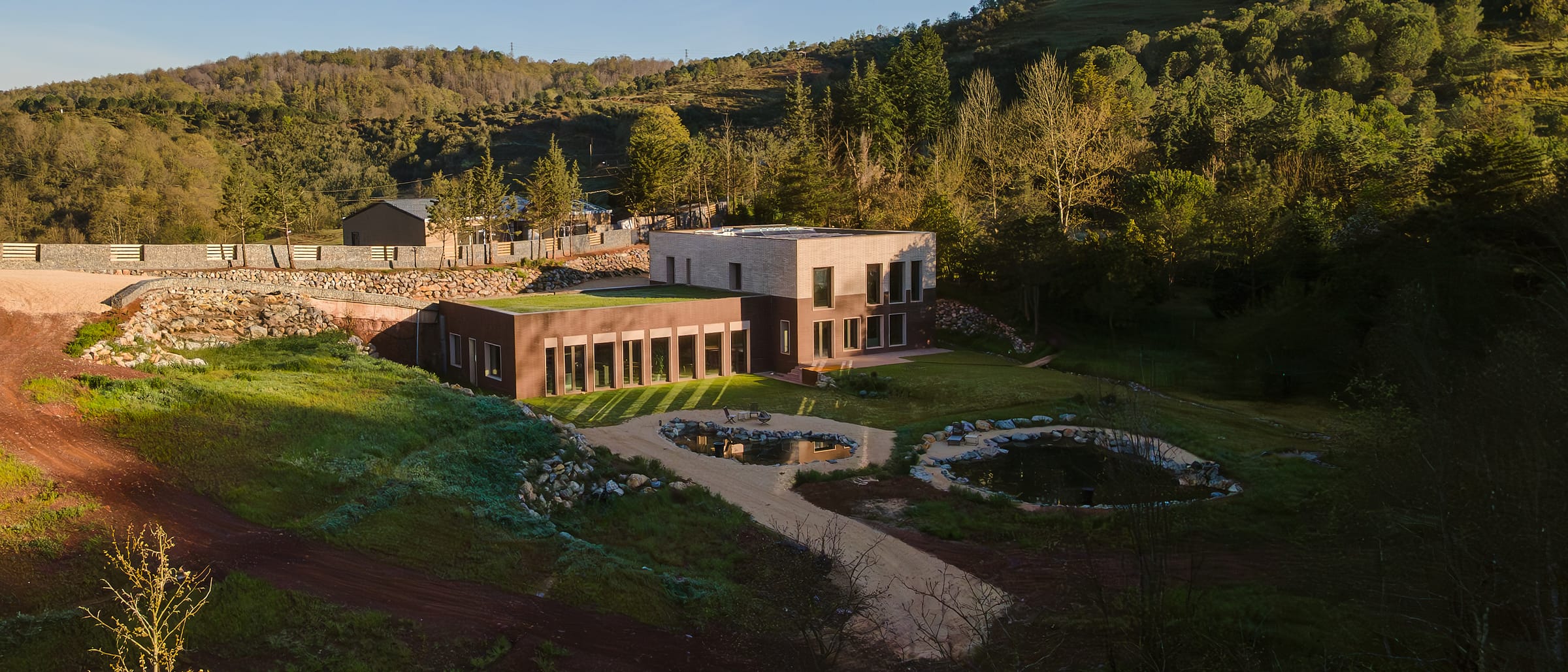 Aerial view of a long low brick building set into a green slope, with two ponds and paths below it.