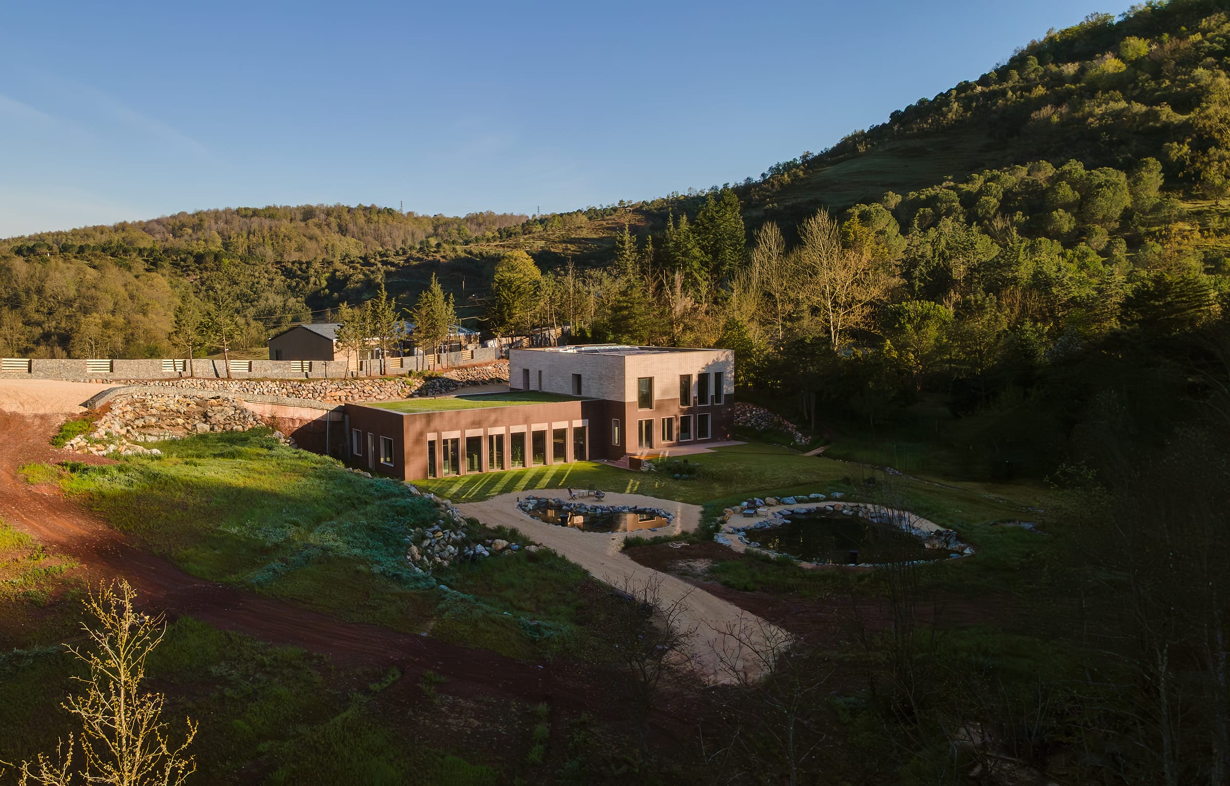 Aerial view of a long low brick building set into a green slope, with two ponds and paths below it.