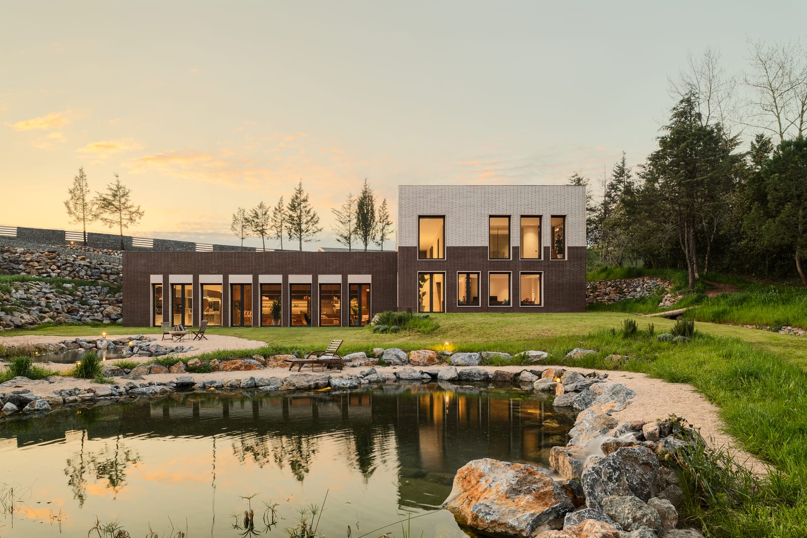 Atlı House at dusk seen across a rock-edged pond, its windows lit warm against a pale evening sky.