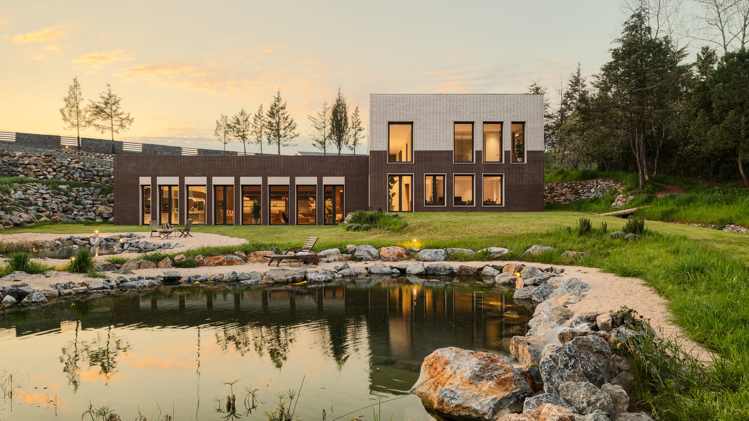 Atlı House at dusk seen across a rock-edged pond, its windows lit warm against a pale evening sky.